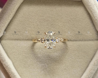 May include: A gold ring featuring a central oval-cut stone flanked by two smaller round-cut stones on each side. The band is thin and polished, holding the stones in a prong setting. The ring is centered within a light gray, hexagonal velvet box.
