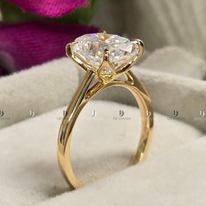 May include: A gold ring with a large, clear, round-cut gemstone. The ring has a delicate, four-prong setting and a smooth, polished band. The gemstone sparkles, reflecting light. The ring is presented on a soft, white surface.