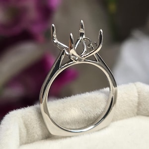 May include: A silver ring setting with a pronged design to hold a gemstone. The ring has a square band and a detailed setting, showcasing a modern and elegant style. The ring is displayed on a white surface.