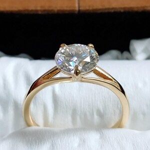 Op de afbeelding: Een gouden verlovingsring met een grote, ronde, heldere edelsteen. De ring heeft een klassieke vier-klauwzetting en een eenvoudige, gepolijste gouden band. De ring wordt gepresenteerd in een bruine fluwelen ringdoos met een witte satijnen binnenkant.