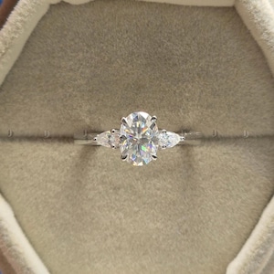 May include: A silver ring with a large oval-cut diamond center stone, flanked by two smaller pear-shaped diamonds. The ring is set in a light-colored velvet ring box, showcasing the sparkling gemstones.