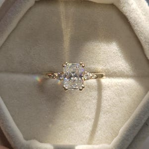 Puede incluir: Un anillo de compromiso dorado con una gran piedra preciosa rectangular y transparente como pieza central. El anillo tiene piedras de acento más pequeñas a cada lado de la piedra principal. El anillo está en un joyero de terciopelo.