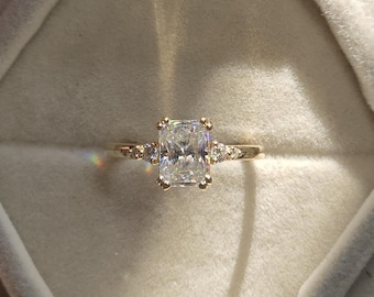 May include: A gold engagement ring with a large, rectangular, clear gemstone as the centerpiece. The ring has smaller accent stones on either side of the main stone. The ring is set in a velvet ring box.