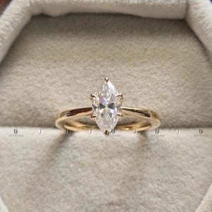 May include: A gold ring with a marquise-cut diamond. The diamond is set in a four-prong setting and the band is a simple, polished gold. The ring is displayed in a gray velvet ring box.