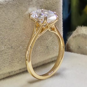 Puede incluir: Un anillo de oro con una gran piedra preciosa transparente de corte ovalado, sostenida por un engaste de garras elevado. La banda es lisa y pulida, mostrando un diseño clásico con una piedra facetada prominente que refleja la luz desde su superficie.