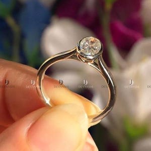 May include: A gold-colored ring with a round, clear gemstone. The ring is held in a person's hand, with a blurred floral background. The gemstone is the focal point, set in a bezel setting.
