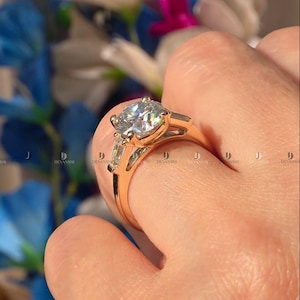 May include: A rose gold ring featuring a large round brilliant-cut center stone held by four prongs, flanked by two smaller baguette-cut side stones. The band is polished rose gold, showcasing a classic three-stone design with clear, faceted gemstones.