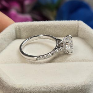 May include: A silver engagement ring with a large, oval-cut diamond center stone. The band is adorned with smaller diamonds, and the ring is marked "18KT". The ring is set in a light gray velvet box.
