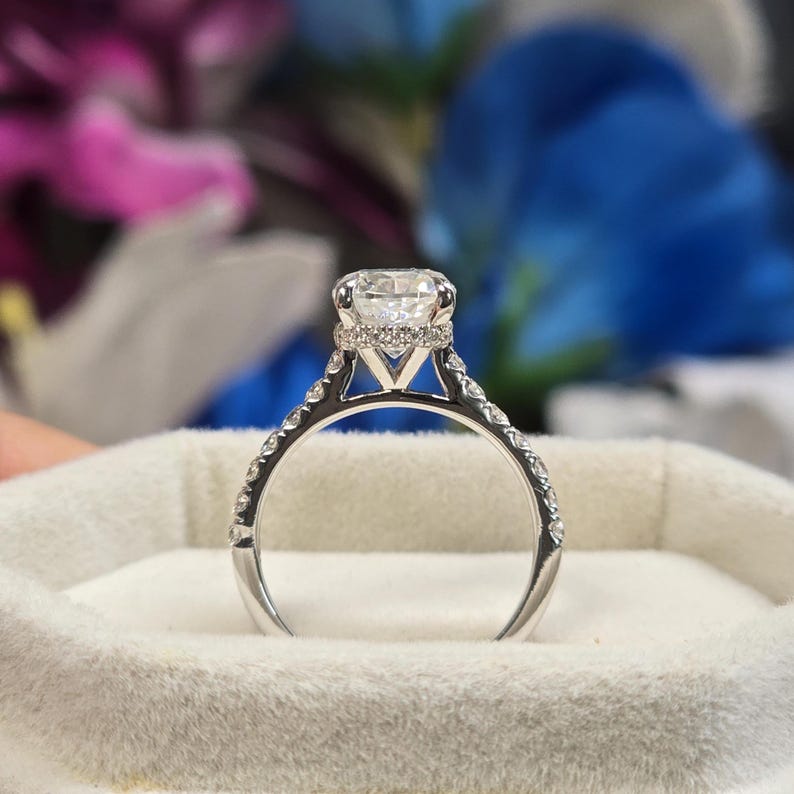 May include: A silver engagement ring featuring a large, round, clear diamond. The band is set with smaller diamonds. The ring is presented in a white velvet box. The design is elegant and classic, suitable for a proposal.