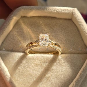 Può includere: Un set di anelli di fidanzamento in oro con una pietra centrale di diamante taglio rettangolare e pietre di accento più piccole. La fascia è anch'essa adornata da diamanti più piccoli. L'anello è presentato in un portagioie di colore chiaro.