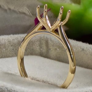 May include: A gold-colored ring setting with a pronged design to hold a gemstone. The ring has a polished finish and a classic, elegant style. The prongs are curved and extend upwards.