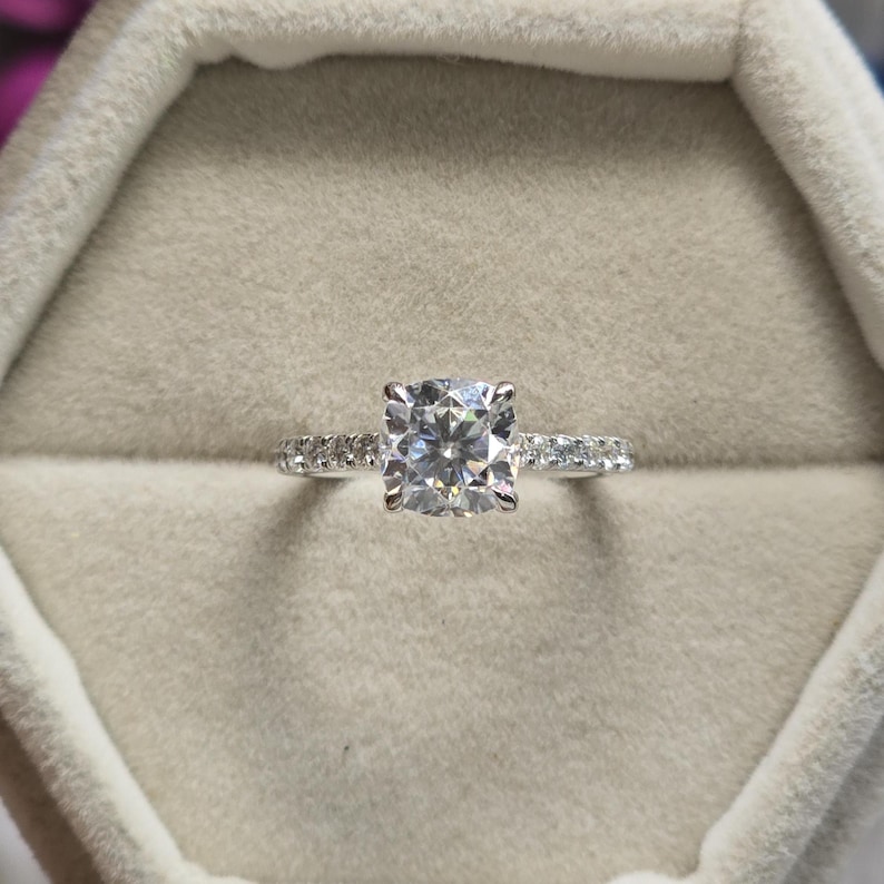 May include: A silver ring with a large, square-cut, clear gemstone as the centerpiece. The band is adorned with smaller, clear gemstones. The ring is set against a soft, neutral-toned background, highlighting its sparkle and design.