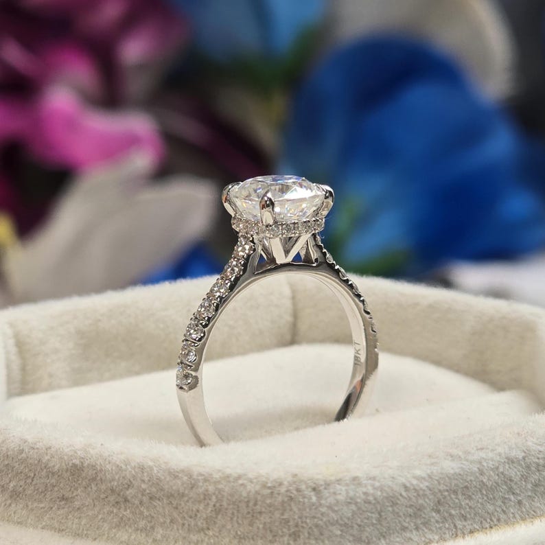 May include: A close-up of a diamond engagement ring. The ring features a large, round-cut diamond set in a prong setting. The band is made of white metal and is encrusted with smaller diamonds. The ring is displayed in a white velvet box.