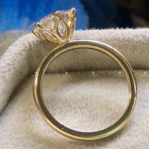Könnte beinhalten: Ein goldener Ring mit einem großen, klaren Edelstein. Der Edelstein ist in einer detaillierten, floral inspirierten Fassung gefasst. Der Ringband ist glatt und poliert. Der Ring wird auf einer neutralfarbenen Oberfläche präsentiert.
