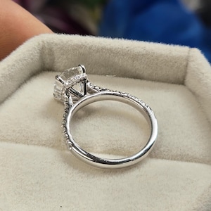 May include: A close-up of a silver engagement ring with a large, square-cut clear gemstone. The band is encrusted with small, sparkling stones. The ring is set against a soft, light gray background.