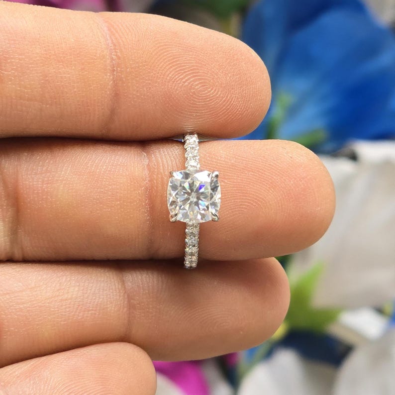 May include: A close-up of a diamond ring with a square-cut center stone. The band is adorned with smaller, sparkling stones. The ring is held between fingers, showcasing its brilliance and design. The ring is a piece of jewelry.