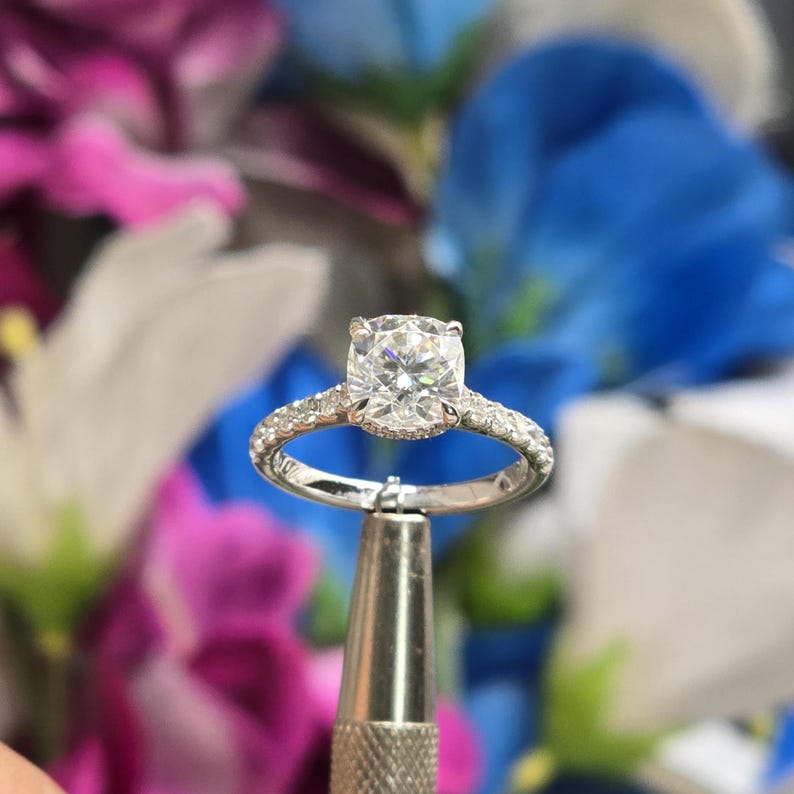 May include: A diamond engagement ring with a large, square-cut center stone. The band is made of white metal and is encrusted with smaller diamonds. The ring is held by a metal tool against a blurred floral background.