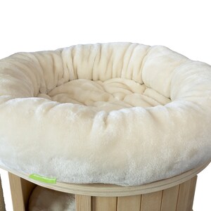 K&ouml;nnte beinhalten: Ein cremefarbenes Katzenbett mit einem weichen, runden Design. Das Bett ist Teil eines mehrst&ouml;ckigen Kratzbaums mit Holzsockel und einer mit Sisal umwickelten Kratzs&auml;ule. Die weiche Textur und die neutrale Farbe bieten einen gem&uuml;tlichen Platz f&uuml;r eine Katze.