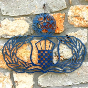 Puede incluir: Adorno de pared de metal azul con un diseño detallado. La pieza incluye un emblema central con un patrón de cuadros, rodeado de hojas de laurel y una esfera decorativa. Montado en una pared de piedra.