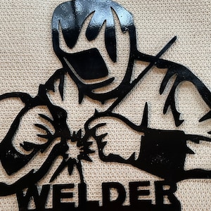 May include: Black metal silhouette of a welder wearing a welding mask and holding a welding torch. The word "WELDER" is written below the silhouette.