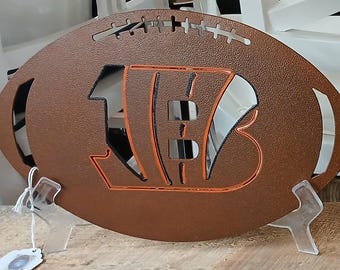 Pode incluir: Uma placa de metal castanho em forma de bola de futebol com o logotipo dos Bengals de Cincinnati recortado em laranja. A placa é exibida em um suporte transparente, mostrando as iniciais da equipe. A placa foi projetada para se assemelhar a uma bola de futebol.