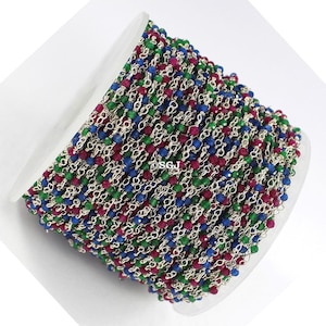 Könnte beinhalten: Eine Spule mit silberner Kette mit roten, grünen und blauen Edelsteinperlen. Die Kette ist um eine weiße Spule gewickelt.