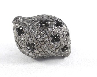May include: A dark gray, oval-shaped bead with a floral pattern. The bead is covered in small, sparkling stones, with dark, openwork floral designs. The bead is set against a white background.