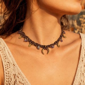 Op de afbeelding: Een bruine macramé choker ketting met een centrale koperen halve maan bedel, afgewisseld met kleine paarse edelsteenkralen, metallic goudkleurige kralen en kleine koperen blaadjesvormige bedels langs de geweven koordstructuur.