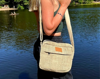 May include: A light beige hemp crossbody bag with a brown leather flap and zipper closure. The bag is worn by a person standing near a lake.