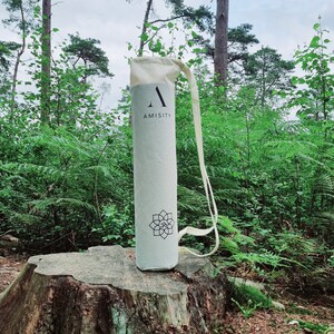 May include: A white canvas yoga mat bag with a black "A" and the word "AMISITY" printed on it. The bag is sitting on a tree stump in a forest setting.