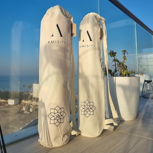 May include: Two white yoga mat bags with black straps and a black logo that says "AMISITY". Each bag has a black Om symbol inside a flower design.