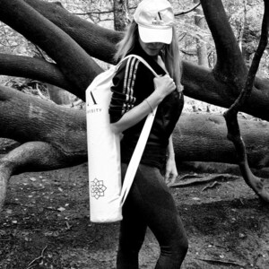 May include: A white yoga mat bag with a black strap and a black and white logo that says "AMISTY" and a symbol. The bag is being carried by a person wearing a black shirt and black pants.