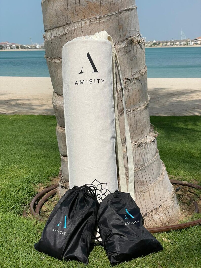May include: Two black drawstring bags with the word "AMISITY" printed on them, and a white canvas bag with the word "AMISITY" printed on it. The bags are sitting on a green grassy surface.