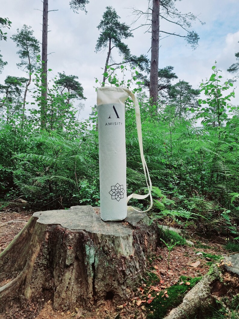 May include: A white canvas yoga mat bag with a black "A" and the word "AMISITY" printed on it. The bag is sitting on a tree stump in a forest setting.