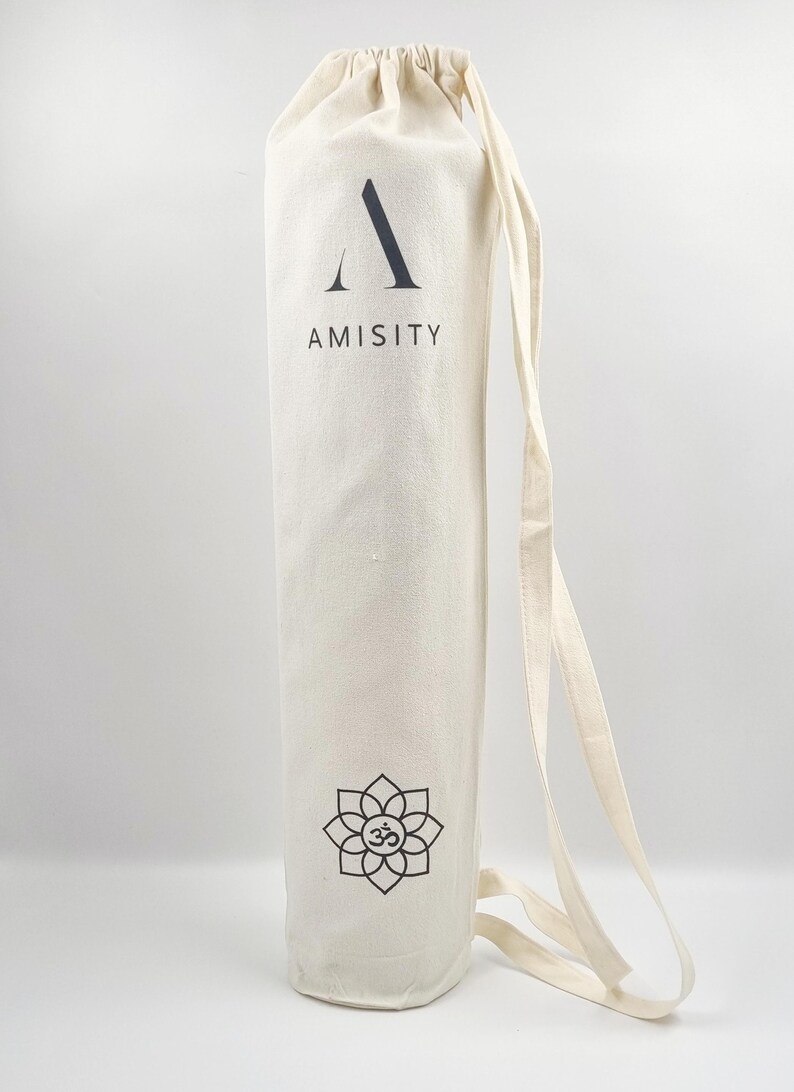 May include: A white canvas yoga mat bag with a black drawstring closure. The bag has a black logo that says "AMISITY" and a black lotus flower with an "OM" symbol in the center.