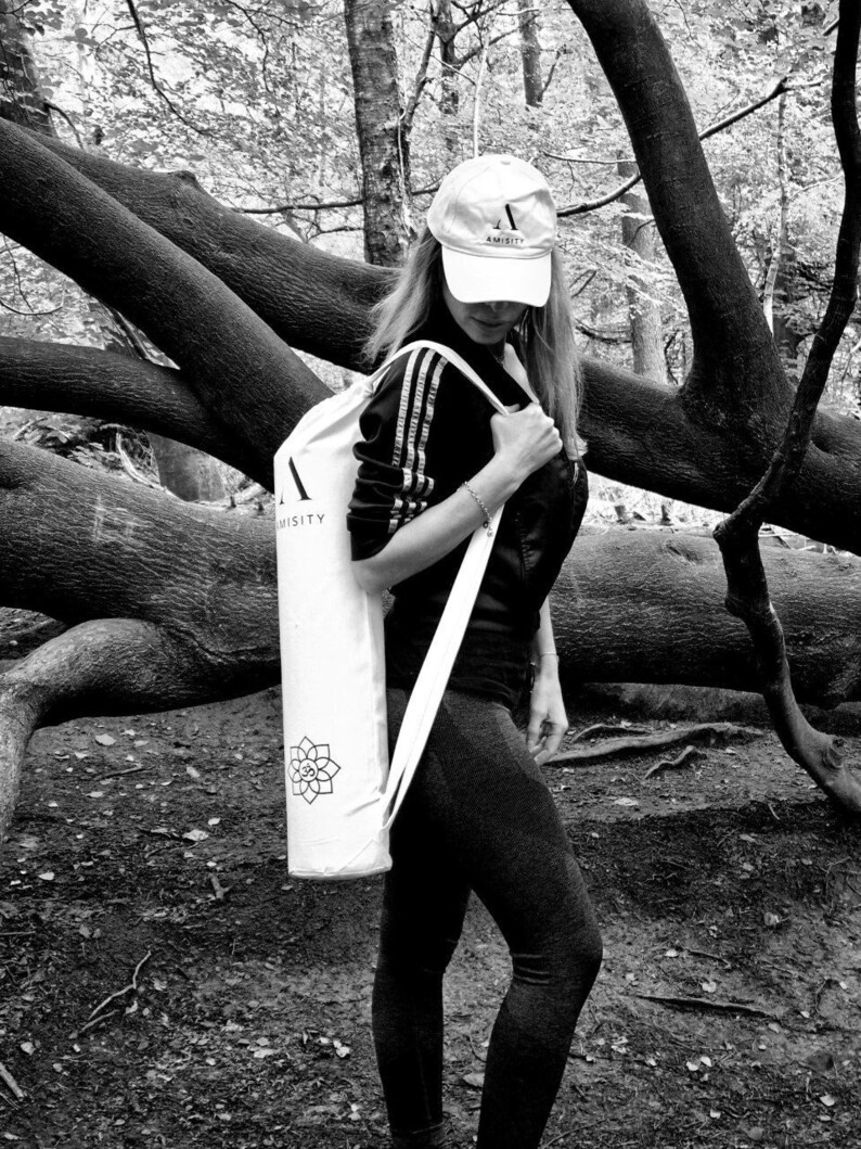 May include: A white yoga mat bag with a black strap and a black and white logo that says "AMISTY" and a symbol. The bag is being carried by a person wearing a black shirt and black pants.