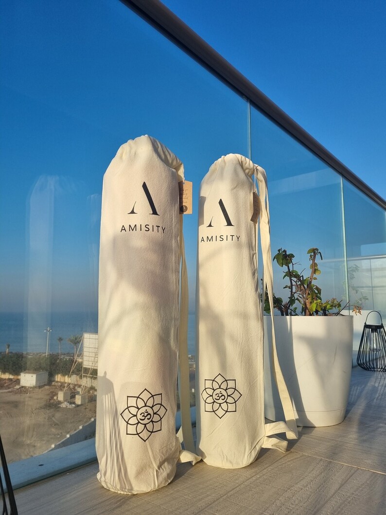 May include: Two white yoga mat bags with black straps and a black logo that says "AMISITY". Each bag has a black Om symbol inside a flower design.