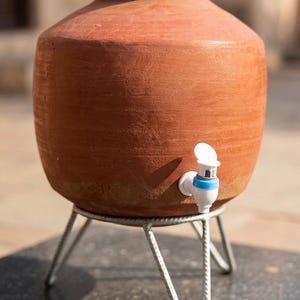 Könnte beinhalten: Ein Terrakotta-Wasserspender mit einem weißen und blauen Wasserhahn, auf einem silbernen Metallständer. Der Tontopf hat eine abgerundete Form und eine weite Öffnung. Der Spender ist für den Außenbereich konzipiert.