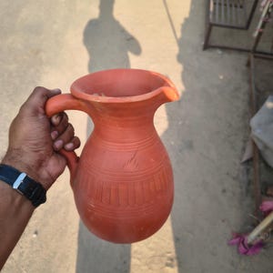 May include: A terracotta jug with a handle and a spout. The jug has a rounded body with etched vertical lines and horizontal bands. The jug is held by a person.