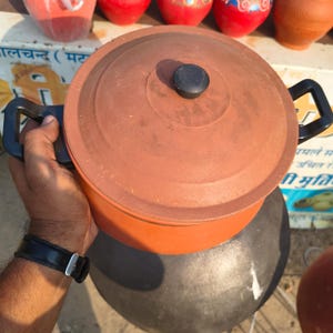 May include: A terracotta cooking pot with a matching lid and black handle. The pot is round and has a dark brown color. The lid has a black knob. Other terracotta pots are visible in the background.