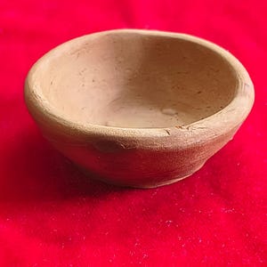 May include: A small, round, unglazed terracotta bowl. The bowl has a simple, smooth design and a light brown color. It sits on a red surface.