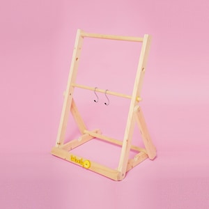 Può includere: Un attaccapanni in legno colorato chiaro con due ganci argentati appesi a una barra orizzontale. L'attaccapanni ha una base rettangolare e un design a forma di A. La parola "World" è stampata sulla base con una faccina gialla.