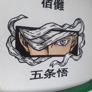 K&ouml;nnte beinhalten: Gestickter Aufn&auml;her mit einem stilisierten Anime-Gesicht mit blauen Augen und wei&szlig;en Haaren. Das Design enth&auml;lt japanischen Text &uuml;ber und unter dem Gesicht, alles auf wei&szlig;em Hintergrund.