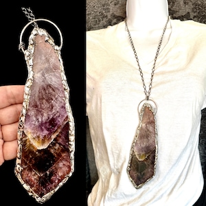 Może przedstawiać: Ręcznie robiony naszyjnik z wisiorkiem z ametystu. Duży, nieregularny kamień ametystu jest osadzony w srebrnej ramie. Wisiorek zawieszony jest na długim, srebrnym łańcuszku. Kamień ma odcienie fioletu, brązu i żółci.