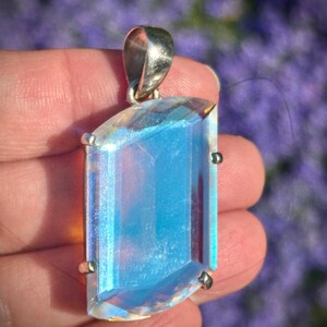 May include: A rectangular pendant featuring a faceted, iridescent blue stone set in a silver-toned metal prong setting. The stone displays a translucent quality with a shimmering, light-reflecting surface and a polished metallic bail at the top.