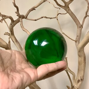 Puede incluir: Una esfera de vidrio verde esmeralda brillante. La superficie lisa y reflectante de la esfera captura la luz, creando reflejos brillantes. La esfera se sostiene en una mano, con un fondo de ramas de árboles desnudas de color claro.
