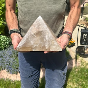 May include: A large, translucent, amber-colored crystal pyramid. The pyramid is held by a person wearing a green shirt and blue shorts. The crystal has a natural, textured appearance and is displayed outdoors in natural light.