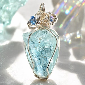 May include: A handcrafted pendant featuring a light blue, irregularly shaped stone wrapped in silver-colored wire. The pendant is adorned with small blue crystals and silver beads at the top, creating a unique and elegant jewelry piece.