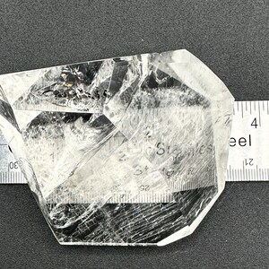 May include: A clear quartz crystal with internal fractures and inclusions, resting on a dark surface. A metal ruler with inch markings is placed next to the crystal for scale. The crystal has a geometric shape.