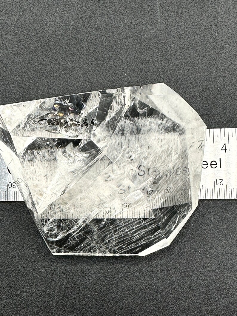 May include: A clear quartz crystal with internal fractures and inclusions, resting on a dark surface. A metal ruler with inch markings is placed next to the crystal for scale. The crystal has a geometric shape.